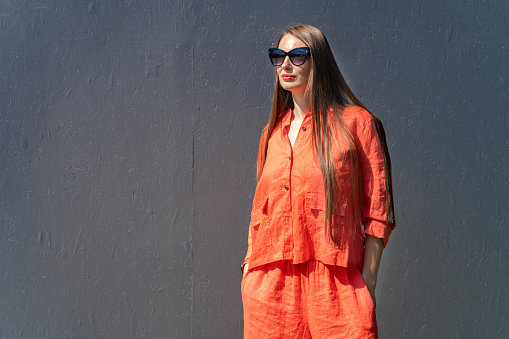 Summer portrait of a girl with long blond hair. A girl in orange clothes and sunglasses stands near a black wall.