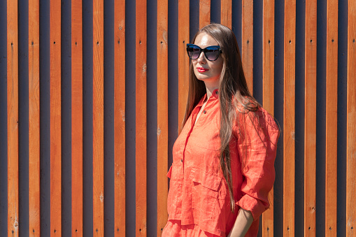 Summer portrait of a girl with long blond hair. A girl in orange clothes and sunglasses stands near a wall of wooden slats.