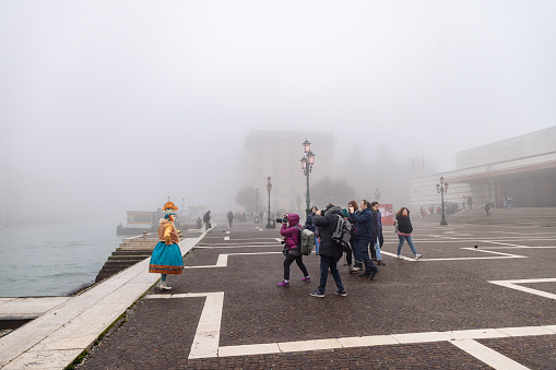 Venice, Italy - February 16 2023: Square by the Grand Canal of Venice, tourists taking photos of mask taking part in the Carnival parade, people walking by on a foggy day at wintertime, Veneto, Europe