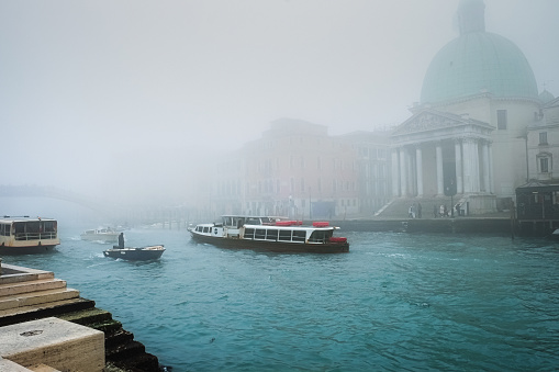 View at special boat vaporetto floating on the water with people commuting down the Grand Canal of Venice on a foggy day in winter, Santa Maria della Salute church in the background