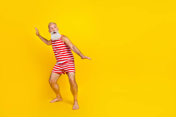 foto de cuerpo entero de un hombre mayor que usa un traje de baño a rayas surfeando manteniendo el equilibrio en el océano mira un espacio vacío aislado sobre un fondo de color amarillo - swimming shorts expressing positivity surfing striped fotografías e imágenes de stock