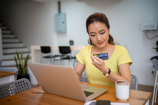 Asian woman sitting and holding credit card for online shopping by laptop and smart phone at home.