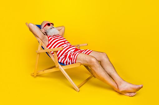 Full length photo of relaxing funky senior man wear red striped lying deck chair hands behind head empty space isolated yellow color background.