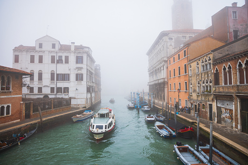 View on canal in Venice with anchored boats along the pier by residential buildings, one boat driving on the water surface, foggy winter day