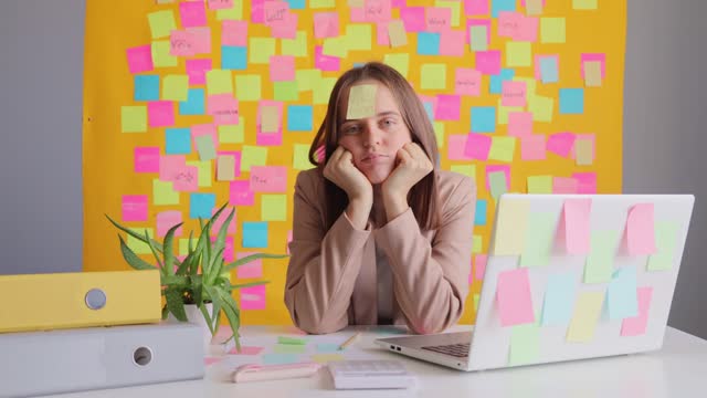 Funny woman freelancer wearing beige jacket sitting in office using laptop computer against yellow wall with colorful memo cards typing on notebook with sticker with vacation inscription on forehead