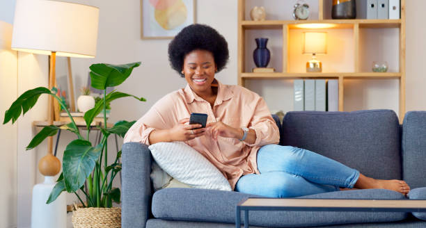 Black woman, smile and living room sofa with phone text and funny web meme at home. Happy, African female person relax and browsing social media on mobile app on the couch with website scroll Laughing woman listening to music and sending a message with funny memes on her phone and drinking coffee while on the sofa. Happy black female browsing social media and surfing the internet online i see you meme stock pictures, royalty-free photos & images