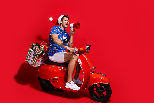 Full size photo of funky young man drive moped megaphone dressed santa claus print x-mas clothes isolated on red color background.
