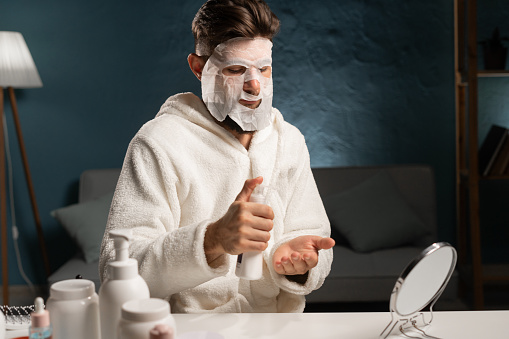 Bearded man with a mask on his face taking skincare squeezing hand cream on palm while sitting in a robe at the dressing table. Copy space