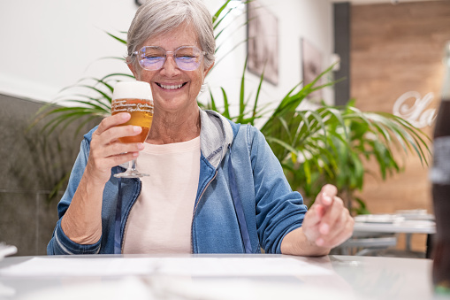 Smiling senior woman sitting at restaurant table with a glass of beer while choosing what to eat from menu