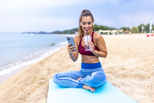 Sporty woman with headphones using her smart phone while relaxing after morning workout at the beach.