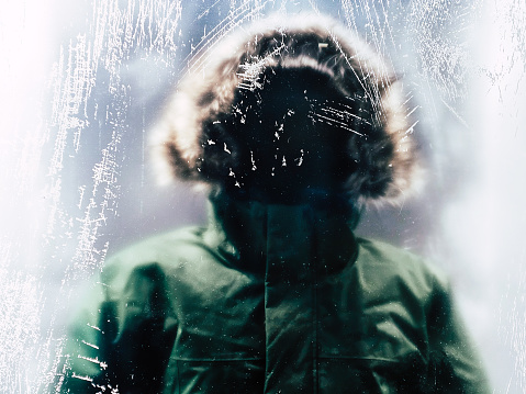 A mysterious, unrecognisable figure wearing a winter parka with fluffy hood stands behind ice and snow-covered glass window.