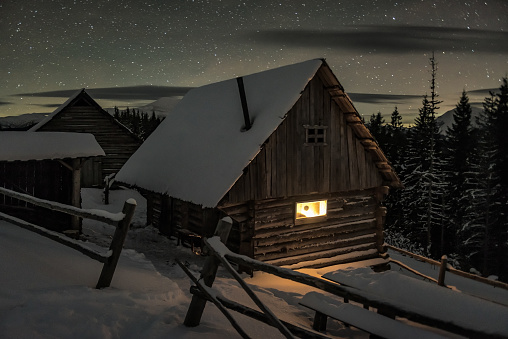 Fantastic winter landscape with glowing wooden cabin in snowy forest. Cozy house in Carpathian mountains. Wide panorama. Christmas holiday concept