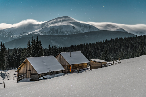 Two wooden log cabins under moonlight in winter Carpathian mountains. Beautiful landscape