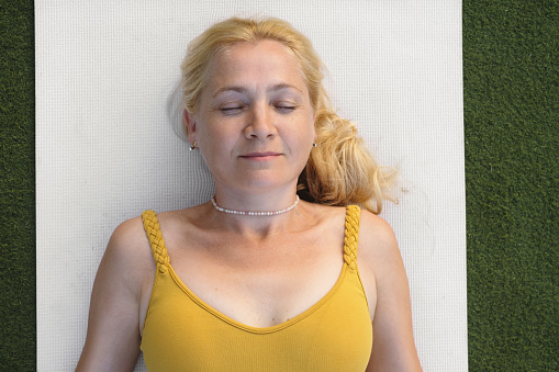 Woman relaxing in shavasana pose after yoga practise. Preventative healthcare for adults. Mindfulness. Female at her 50s laying on mat. Top view