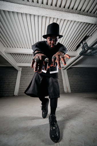 From below full length of serious African American male dancer in extraordinary black outfit and tall hat standing in garage against brick wall and demonstrating hands with accessory