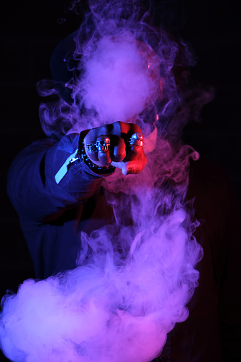 Brutal male model with clenched fist with rings punching through vapor while standing in dark room in studio with pink neon illumination