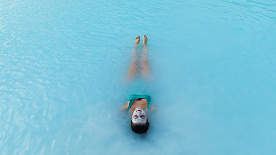 High-angle view of female floating in turquoise hot waters of thermal pool, admiring spa treatment with beauty face mask in Iceland
