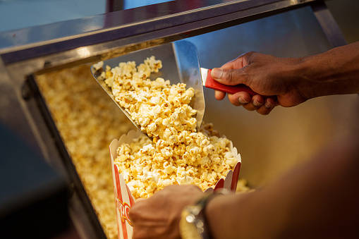 Unidentified person's hand loading popcorn into  bucket at movie theatre concession stand