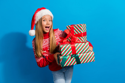Photo of impressed schoolgirl with blond hair dressed red sweater holding gift boxes on new year isolated on blue color background.