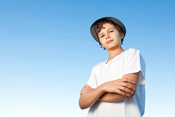 A portrait of a teenage boy, he is standing outside, wearing a white shirt against a blue sky, he is wearing a hat, his arms crossed, looks down into the camera.