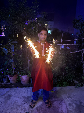 Stock photo of Hindu man dressed in kurta traditional clothing with house decorated with multicolour fairy lights. The man is holding lit sparklers to celebrate Diwali, the Indian traditional festival of lights.