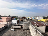 Image of Indian apartment block roof top cityscape, corrugated metal sheets, clothing on clothesline, gas canisters, ladder, air conditioning unit, cloudy blue sky