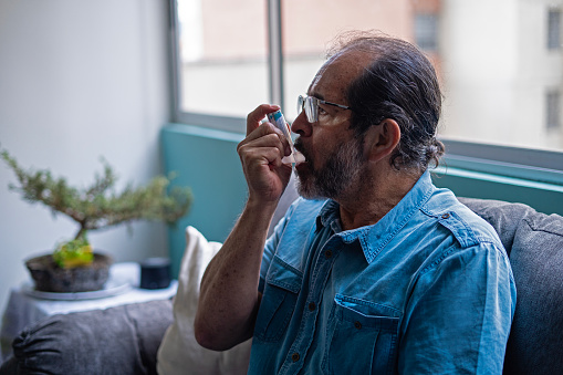 Mature Latino male consciously using an asthma inhaler while at his residence.