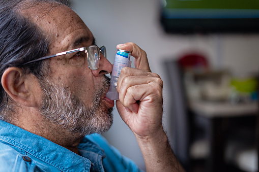Mature Latino male consciously using an asthma inhaler while at his residence.