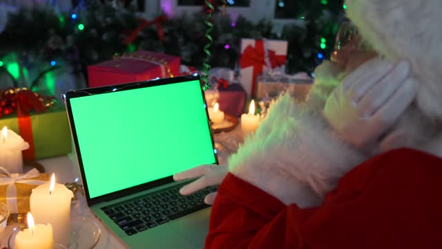 Tired grandfather Santa sits at laptop with chromakey on screen in evening among Christmas decorations