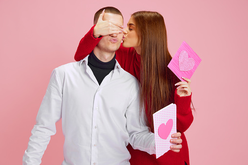 Kiss. Young girl covering boyfriend's eyes and presenting him little gift, postcard with heart against pink studio background. Concept of love, relationship, Valentine's Day, emotions, lifestyle