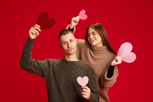 Young happy smiling man and woman, couple, boyfriend and girlfriend posing with paper hearts against red studio background. Concept of love, relationship, Valentine's Day, emotions, lifestyle