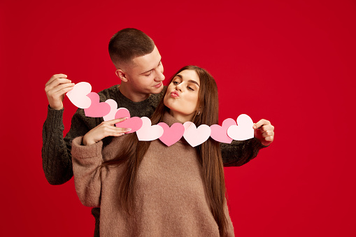 Happy, young couple, boy and girl holding paper heart garland, celebrating holiday against red studio background. Concept of love, relationship, Valentine's Day, emotions, lifestyle