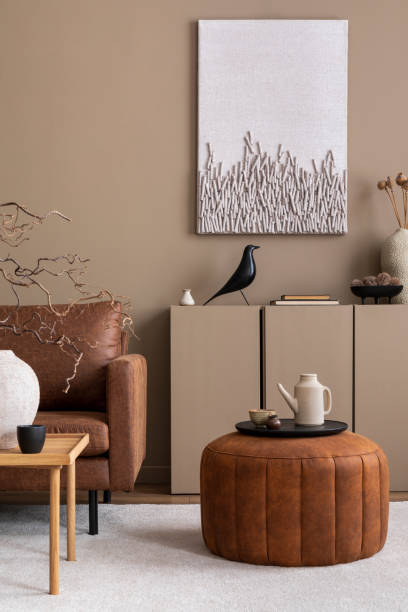 istock Warm and cozy living room interior with mock up poster frame, brown sofa, wooden coffee table, round pouf, beige sideboard, pitcher, cup, books and personal accessories. Home decor. Template. 1809663473