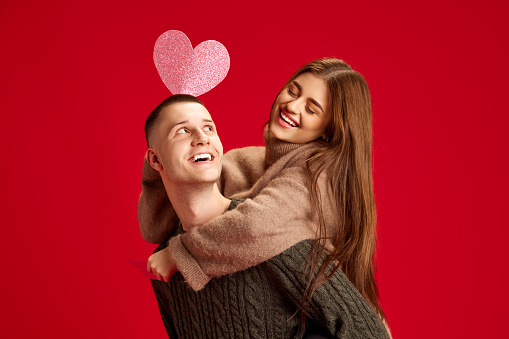 Playful, young, happy couple, man and woman, boyfriend and girlfriend having fun against red studio background. Concept of love, relationship, Valentine's Day, emotions, lifestyle