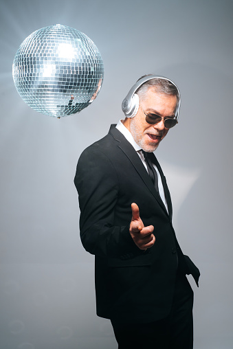 Excited mature businessman in black suit and sunglasses wearing headphones while dancing near shiny disco ball against gray background