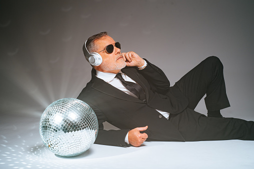 Mature businessman in black suit listening to music through wireless headphones while lying near silver disco ball in gray background