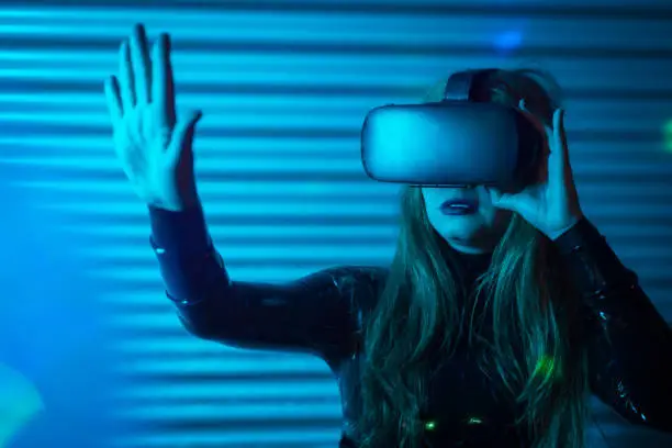 Woman gesturing with the hand while using virtual reality goggles in an urban night space with blue neon light