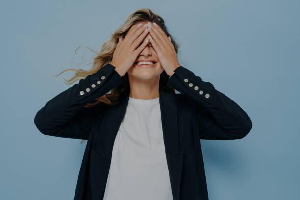 istock Joyful woman in a chic blazer playing 'see no evil' against a cool blue background, embodying carefree spirit and playfulness 1809730517