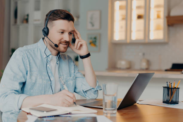 istock Cheerful man with headset engaged in remote work, writing notes beside a laptop in a bright, cozy home setting 1809730522