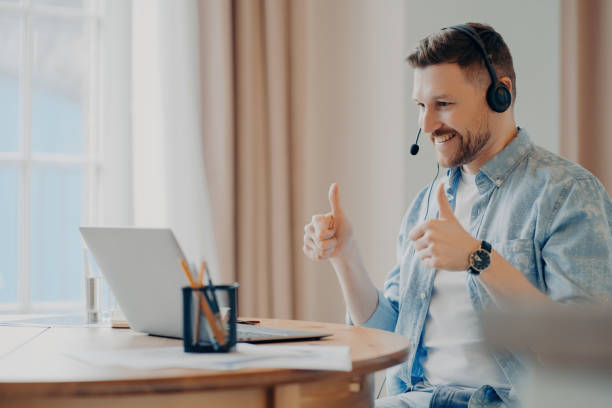 istock Cheerful man giving thumbs up while on a call with a headset, working from a sunny home office with a laptop and stationery 1809730523