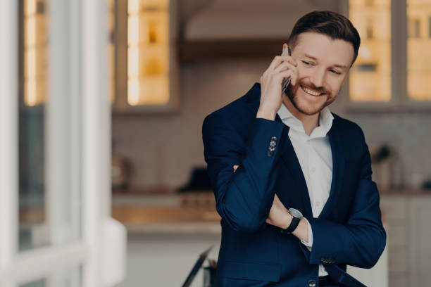istock Confident businessman in a blue suit smiling while talking on the phone, with a modern office backdrop. 1809730525