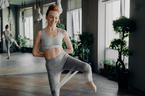 Joyful redhead woman in gym attire gracefully practices resistance band exercises in a sunlit, plant-filled workout space