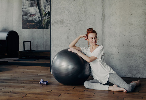 Redhead woman in casual sportswear smiles, relaxing beside a fitness ball in a sunlit workout studio