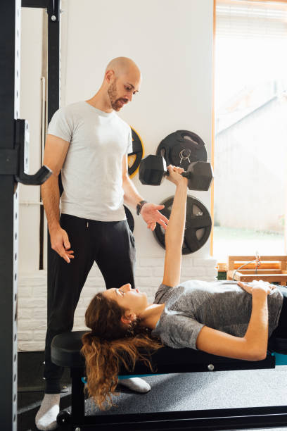 Instructor with young woman lying and doing dumbbell exercise in gym
