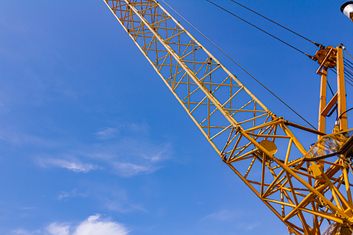 Tall yellow telescopic latticed boom of mobile crane with hoist ropes at the construction site, in background is clear blue sky.