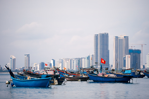 Selective focus on Vietnamese flag on fishing boat moored in port against coast with modern buildings. Da Nang cityscape in Vietnam.