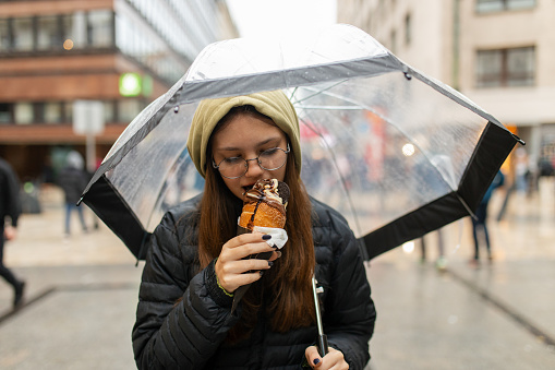 Teenage girl walking in city center, on a rainy day, holding umbrella and enjoys eating Chimney cake in downtown Budapest.