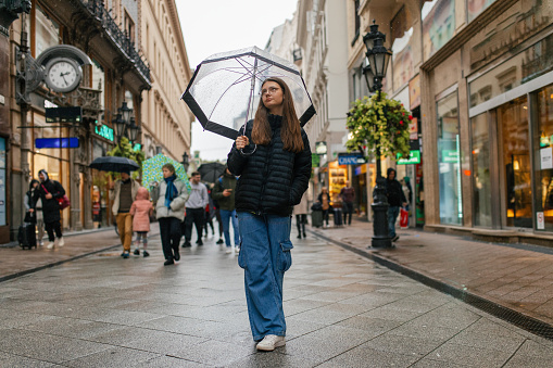 Teenage girl walking in rain in city center in Budapest. She is holding umbrella and looking away from camera.