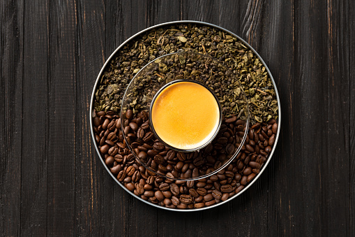 Composition of dry green tea leaves, roasted coffee beans in a round box and coffee in a cup on a dark wooden background top view.