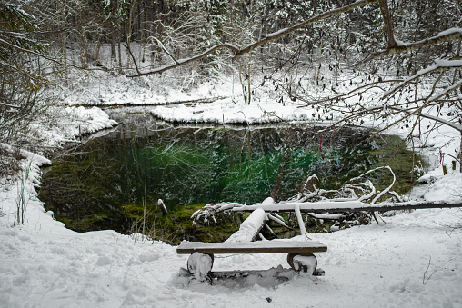 An empty wooden bench covered with snow in front of Blue Springs of Saula (Estonian - Siniallikad) on a cloudy winter day.